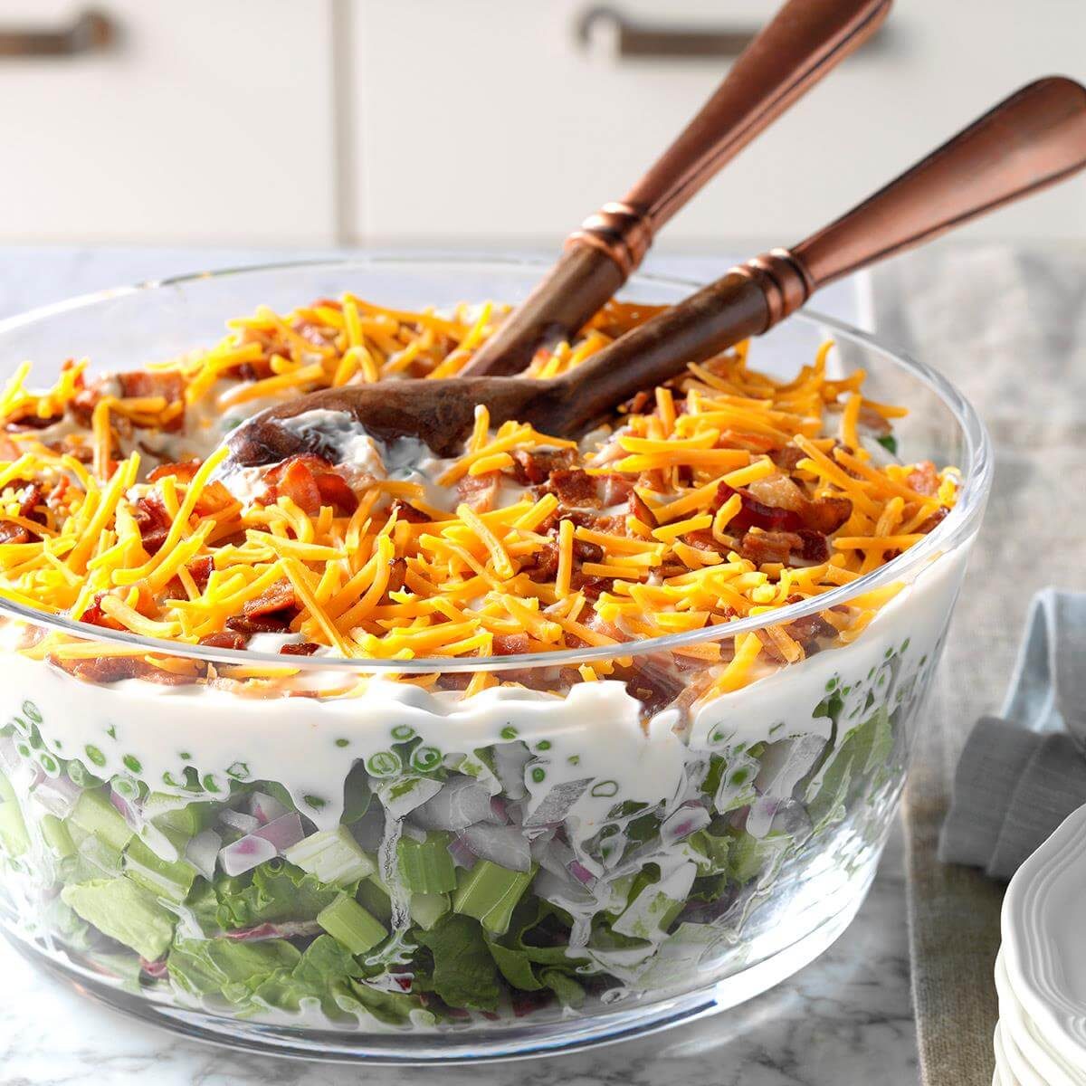 Salad sits in a glass bowl layered with lettuce, onions, mayonnaise, bacon, and cheddar cheese, on a marble countertop with wooden utensils.