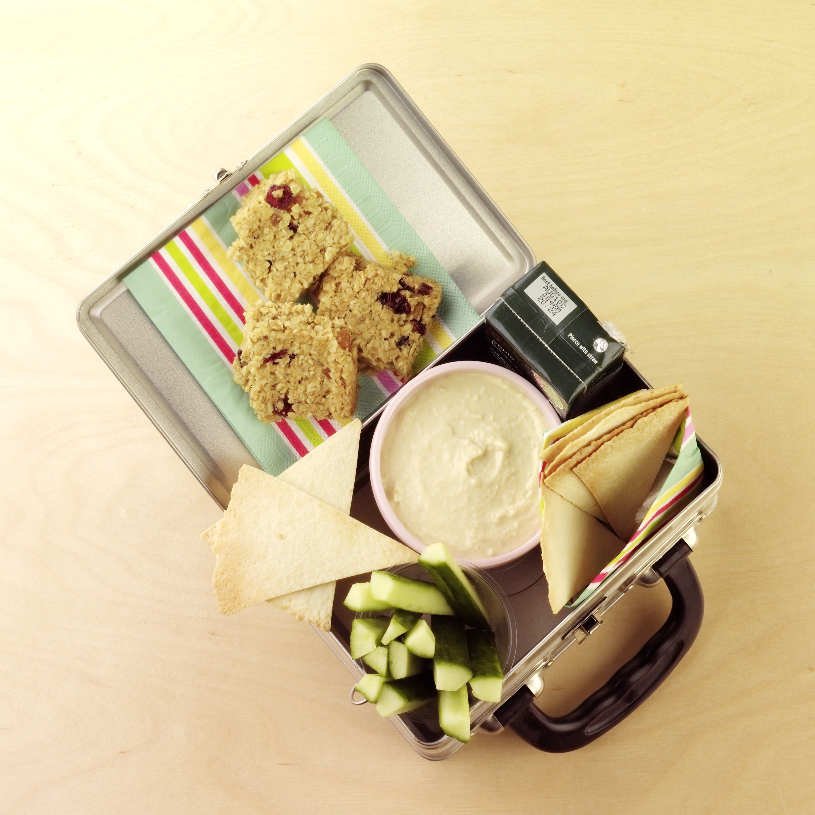 Lunchbox contains energy bars, dip, cucumber sticks, and drink, arranged neatly on colorful napkins on a wooden surface.