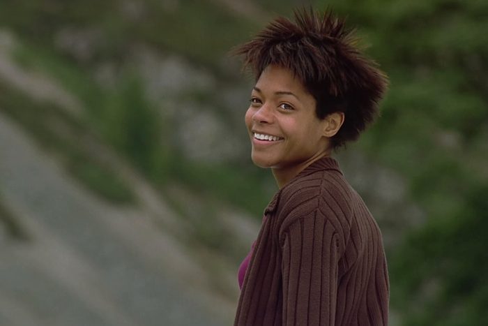 Person smiling while looking back, wearing a brown jacket in a natural, blurred green and gray landscape.
