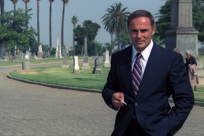 Man in a suit holding a cigarette walks confidently in a cemetery, with gravestones and palm trees in the background.