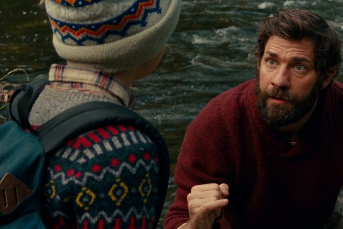 A bearded man wearing a red sweater crouches, looking attentively at a person in colorful winter clothes by a flowing river.