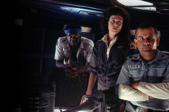 Three people stand intently in a dim, futuristic room, studying equipment. One crouches with hands clasped; two others wear utility jumpsuits.