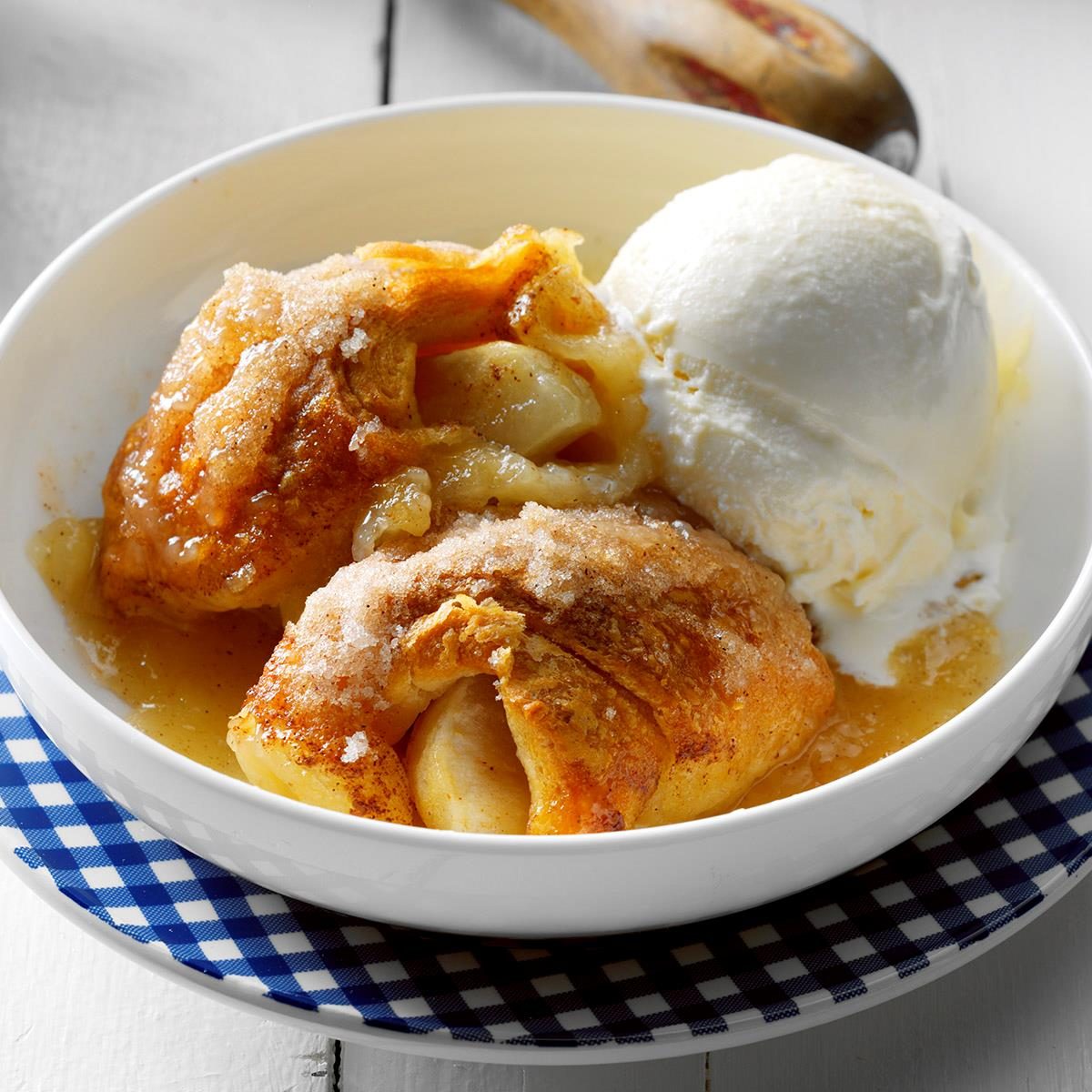 Dish holds apple dumplings, topped with sugar, next to vanilla ice cream; rests on blue-checkered napkin, with a wooden spoon nearby.