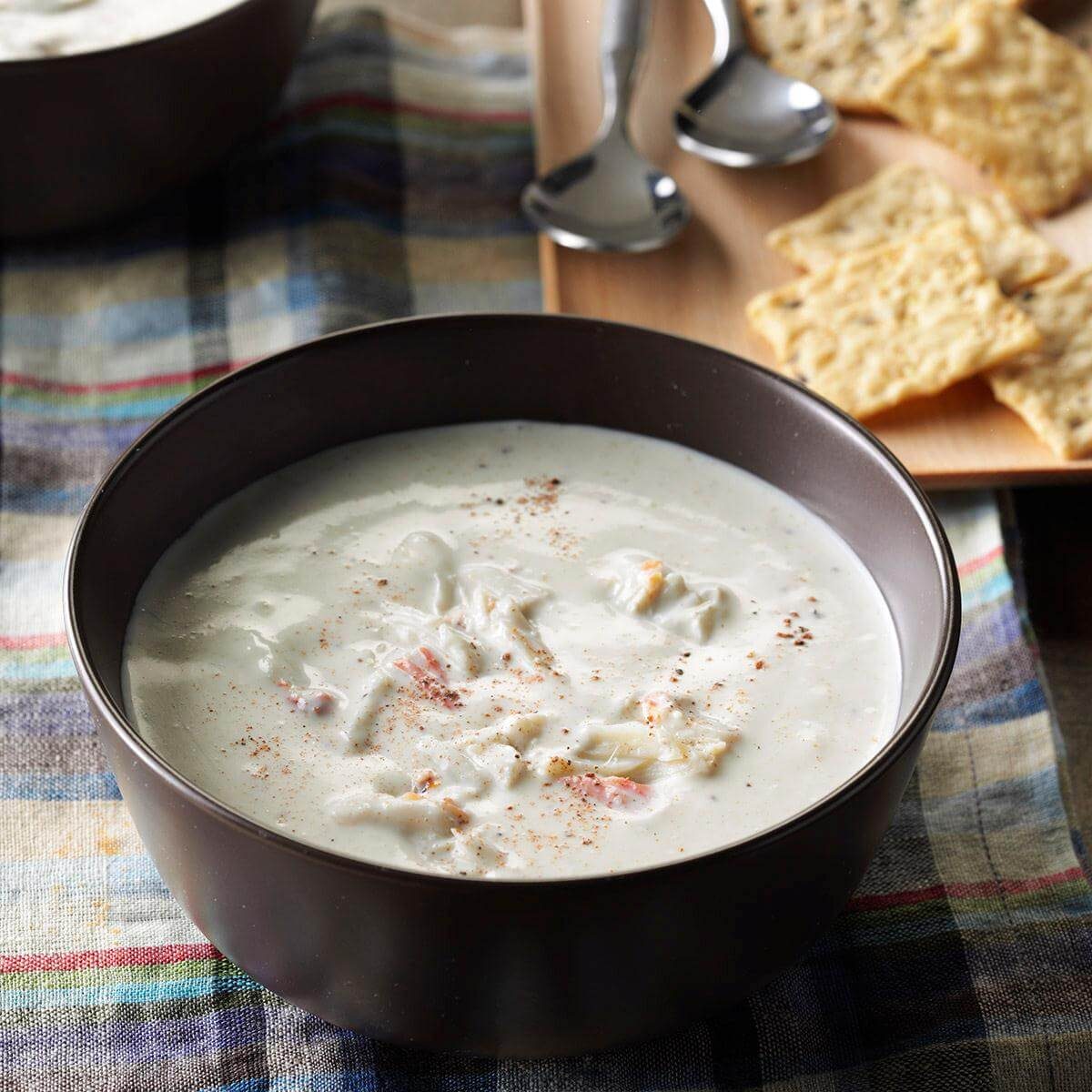A bowl of creamy soup sits on a checkered cloth, accompanied by scattered crackers and two spoons on a wooden tray nearby.