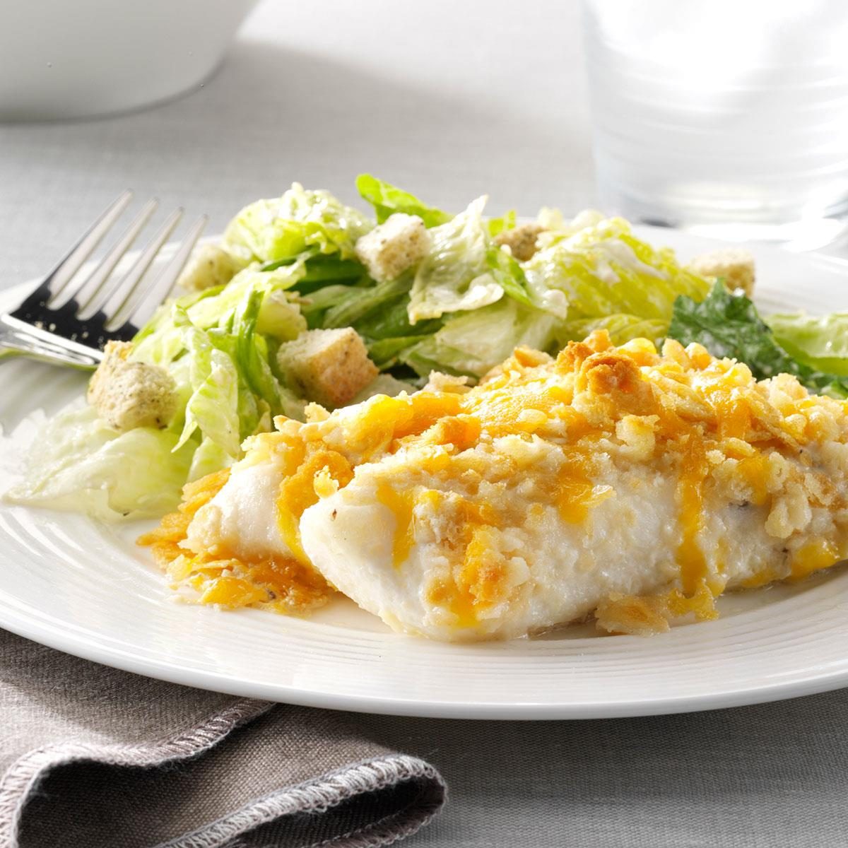 Fish fillet with crispy cheese coating rests beside Caesar salad on a white plate, fork nearby, on a light tablecloth.