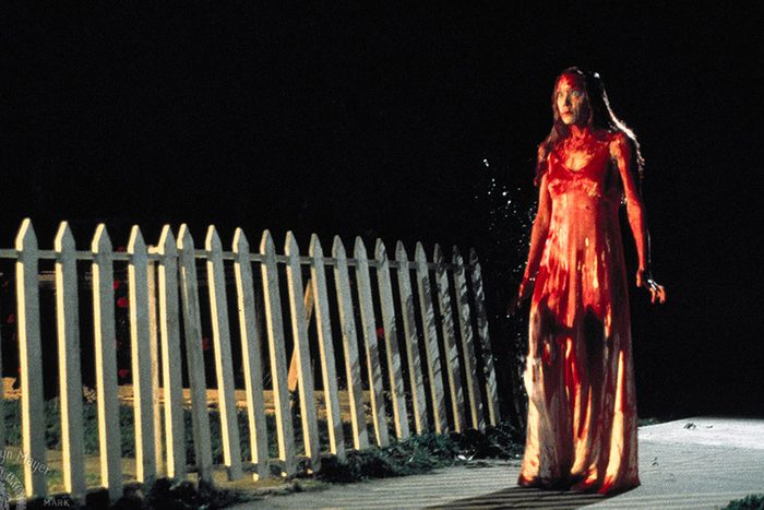 A woman, drenched in a red substance, stands still on a sidewalk at night beside a white picket fence.