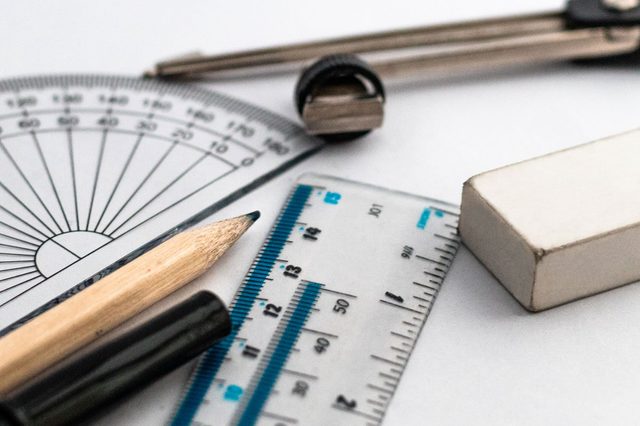 Geometric tools rest on a table; includes a protractor, compass, ruler, pencil, pen, and eraser, arranged for drafting or mathematical work.