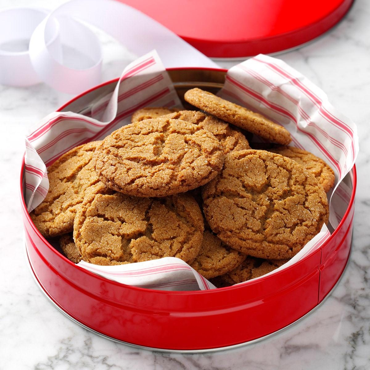 Cookies sit stacked in a red tin, lined with striped paper, on a marble surface, accompanied by a partial view of white ribbon.