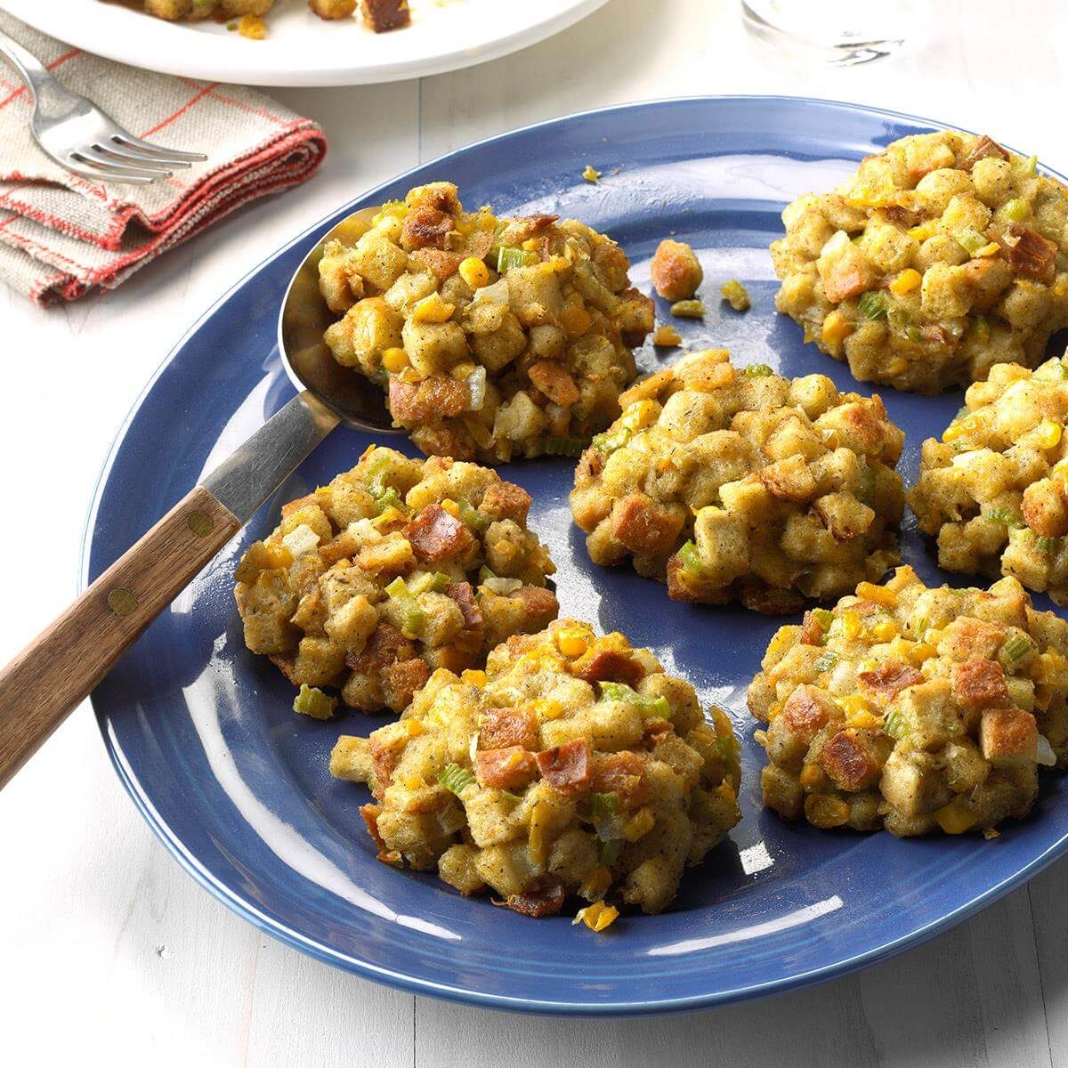 Stuffing balls arranged on a blue plate, ready to be served, with a spoon resting nearby, on a light-colored table with napkins and fork.