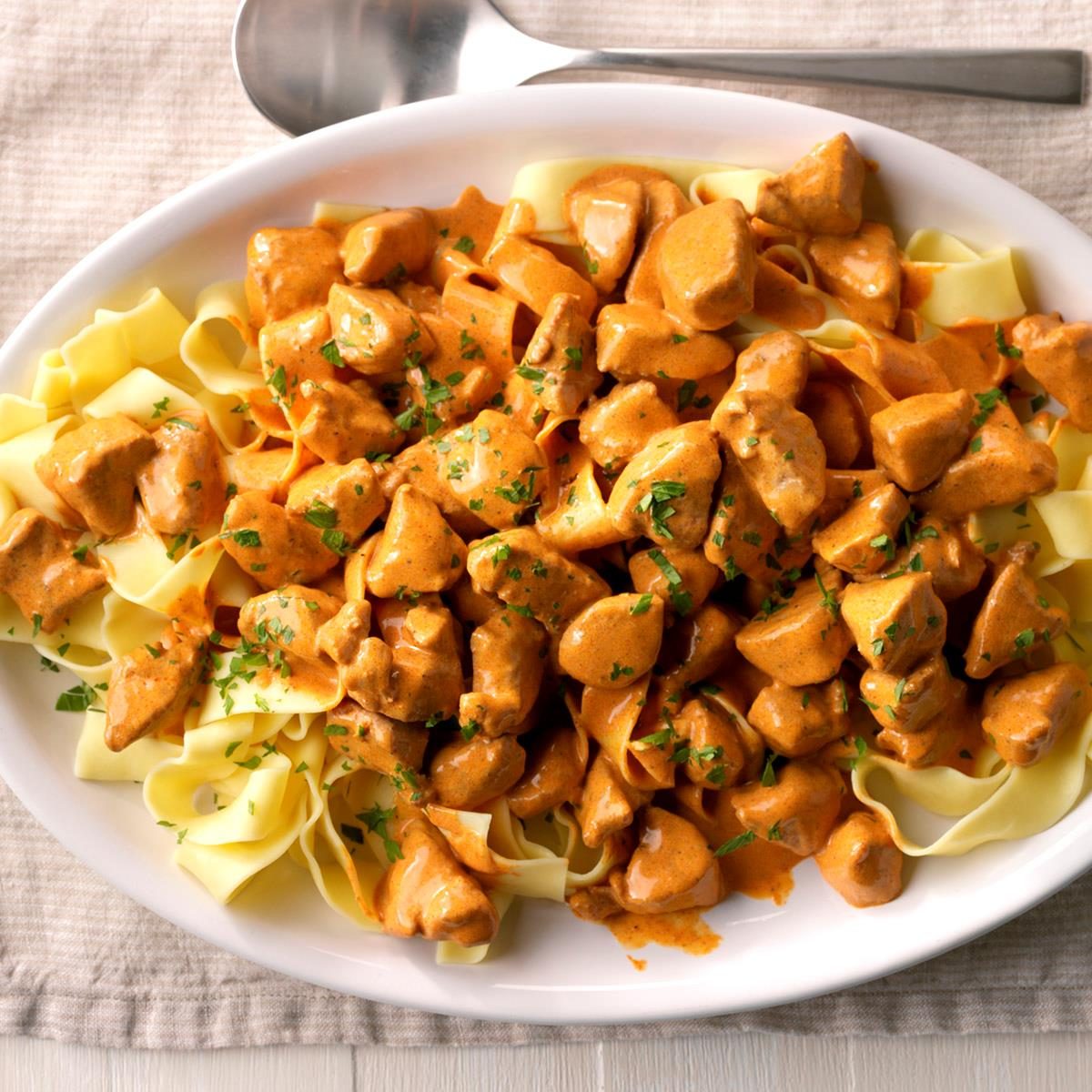 A dish of pasta topped with a creamy, orange sauce and chunks of meat, garnished with chopped herbs, placed on a white plate.