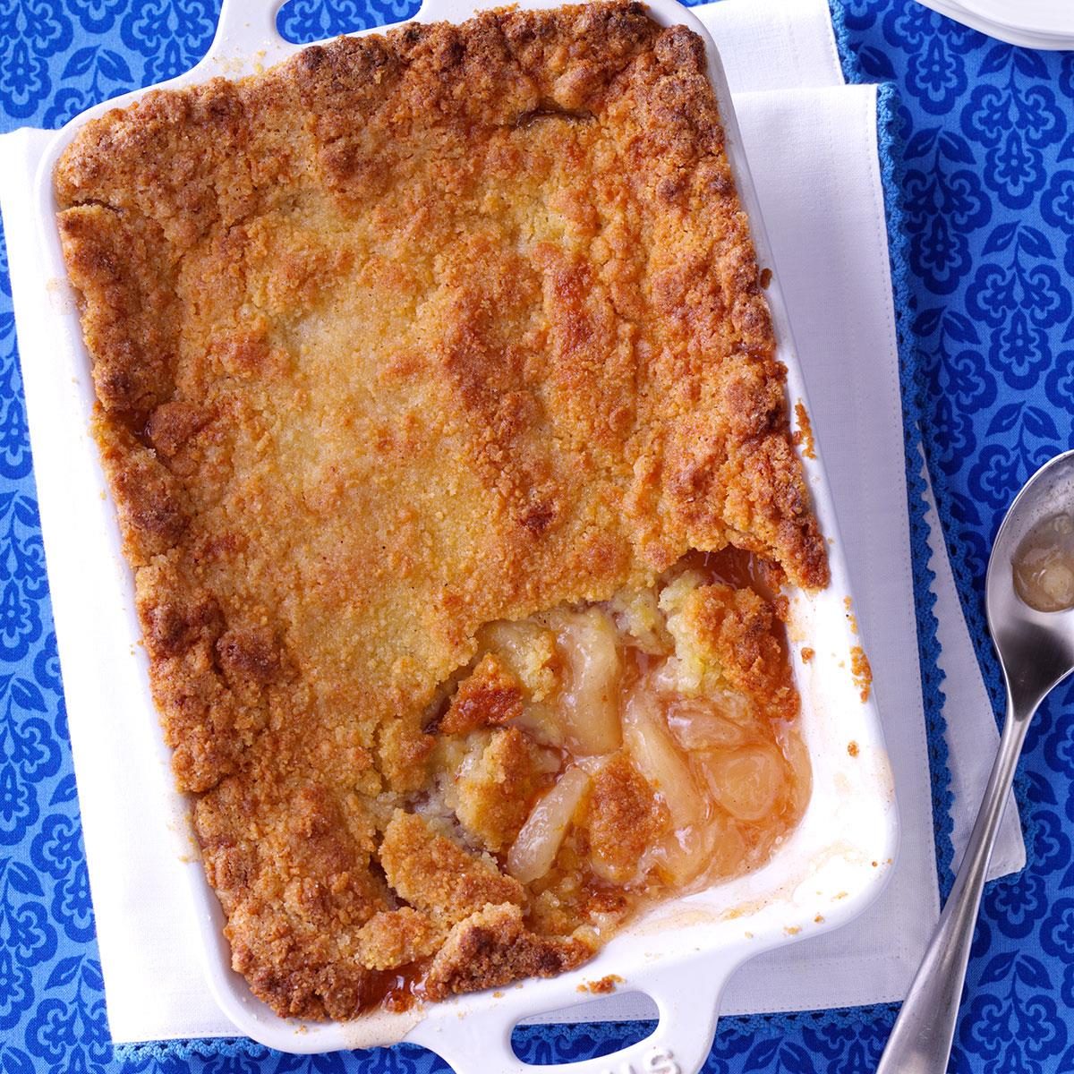 A baked peach cobbler sits in a white dish with a portion missing, revealing juicy peaches, atop a blue patterned tablecloth.