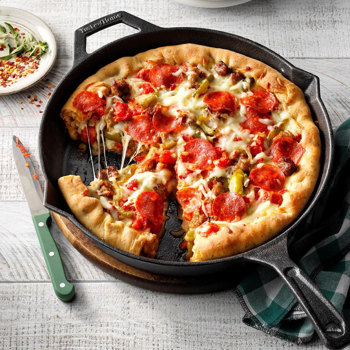 Pizza sits in a cast iron skillet, one slice partially removed, surrounded by a white wooden table, green plaid cloth, and knife.