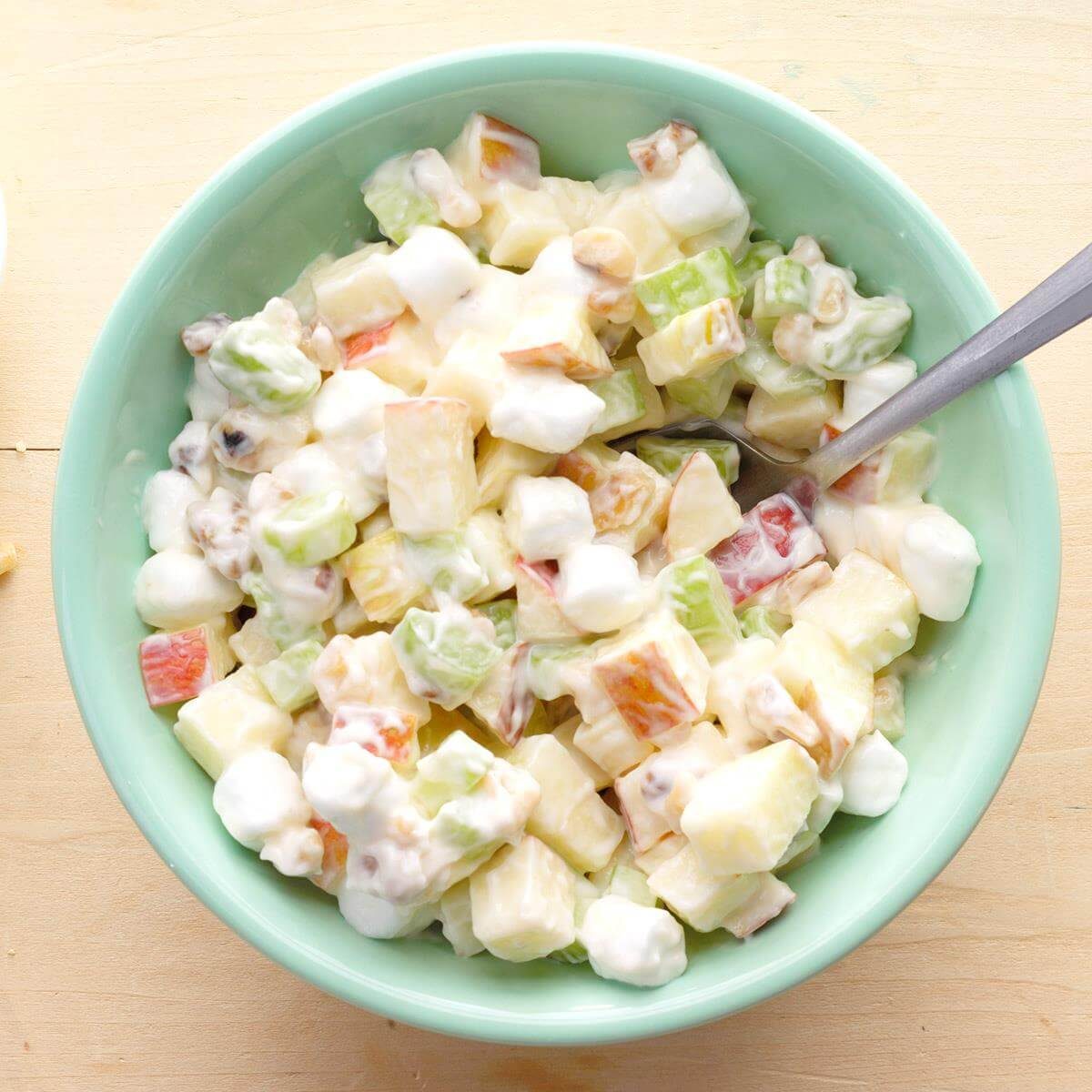 Bowl holds colorful fruit salad mixed with creamy dressing; spoon rests inside. Placed on a light wooden surface.