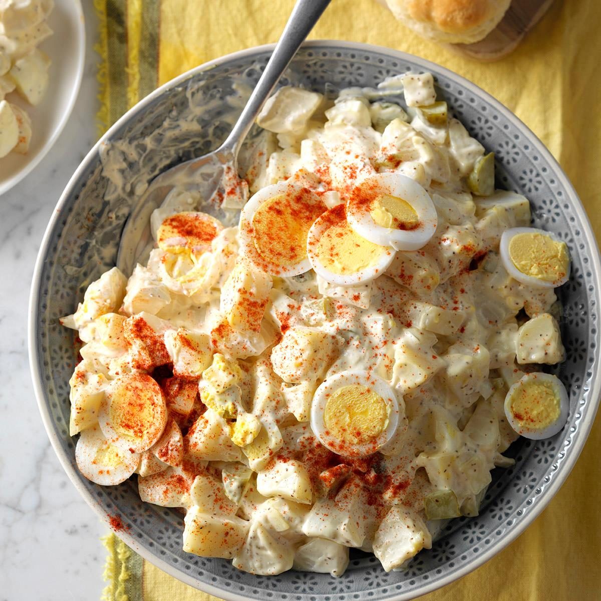 Bowl holds creamy potato salad topped with sliced boiled eggs and paprika; silver spoon rests inside, placed on a yellow cloth.