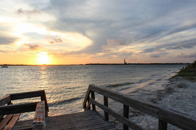 Sunset illuminates the horizon over ocean; wooden boardwalk leads to quiet sandy beach with distant lighthouse under colorful sky.