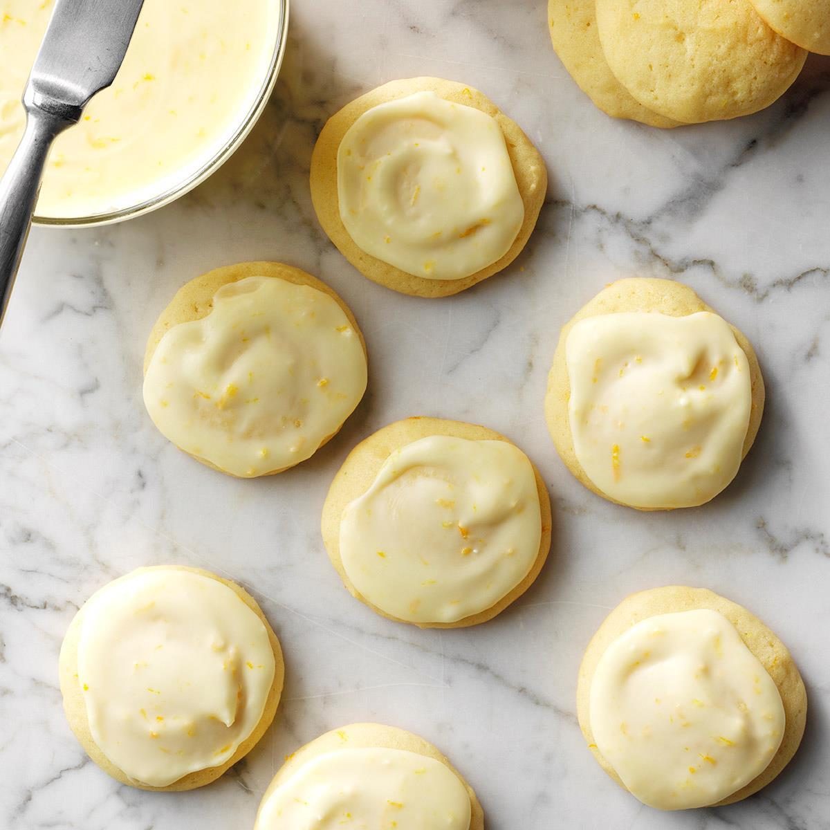 Cookies topped with creamy icing are arranged on a marble surface, next to a bowl of icing and a spatula.