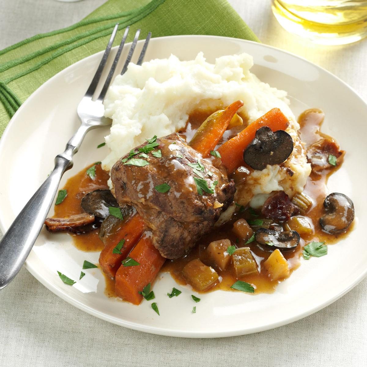 A serving of beef stew sits on a plate with mashed potatoes, carrots, and mushrooms, accompanied by a fork on a napkin.