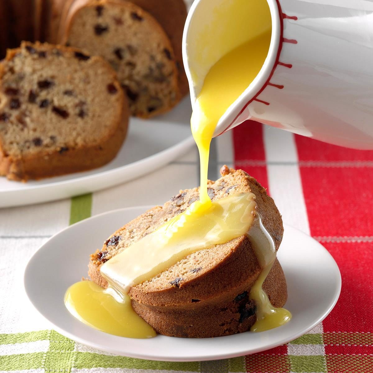 Yellow sauce is poured over two slices of fruitcake on a white plate, set against a plaid tablecloth background.
