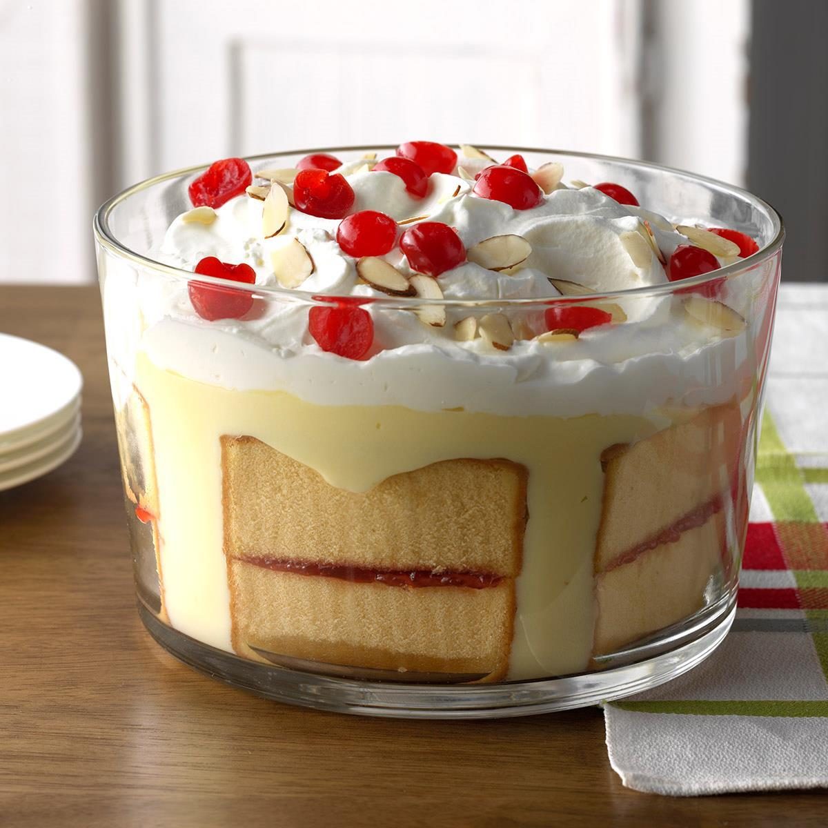 Glass bowl holds layered trifle with cream, cherries, almonds, set on a wooden table with a colorful cloth nearby.