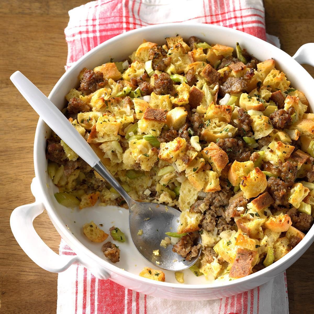 Casserole dish holds baked stuffing with bread cubes, sausage, and vegetables, sitting on a wooden table with a red-and-white checkered cloth.