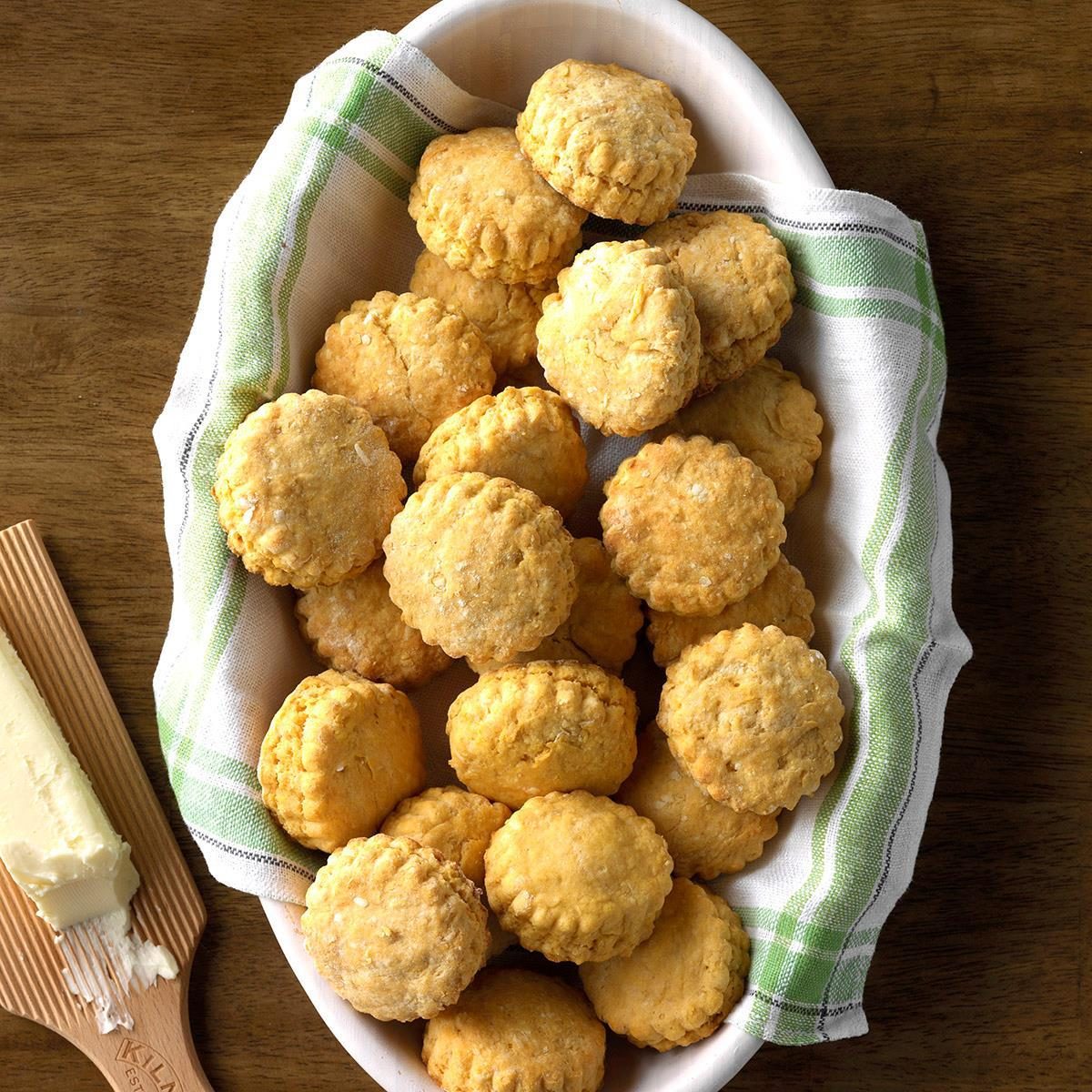 Biscuits sit in a towel-lined basket on a wooden table beside a buttered wooden spreader.