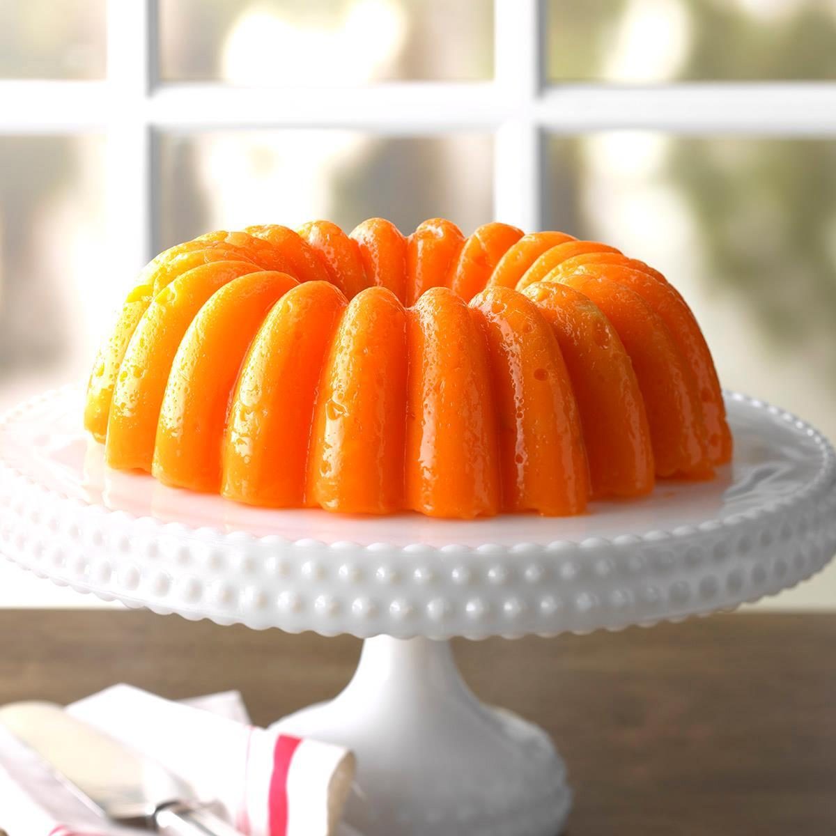 Orange gelatin ring rests on a white cake stand beside cutlery, with a bright window background.