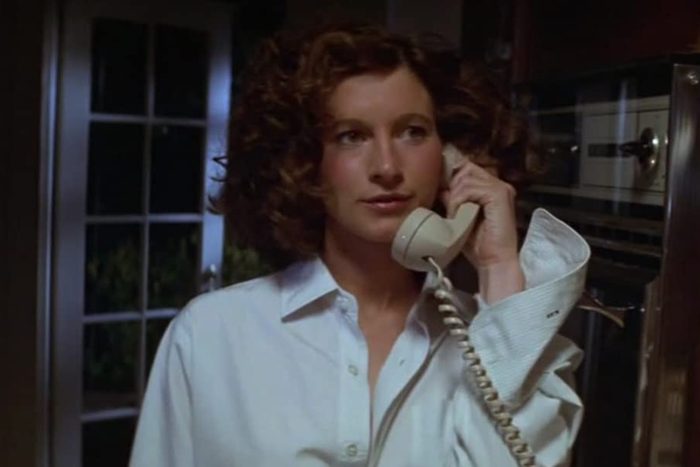 A person holds a corded phone in a domestic kitchen, wearing a white shirt, standing near glass doors.