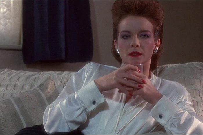 A woman in a white blouse holds a glass, sitting on a sofa in a dimly lit room.