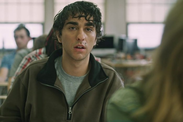 A person looks surprised, seated in a room with blurred individuals and computer screens in the background.