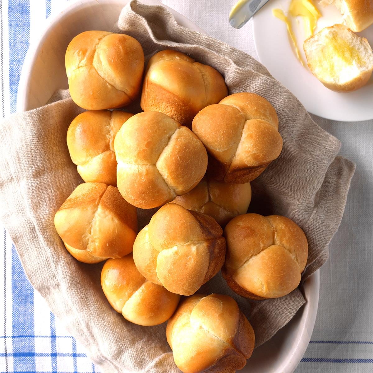 Golden dinner rolls rest in a linen-lined basket, with a buttered roll and knife on a nearby white plate.