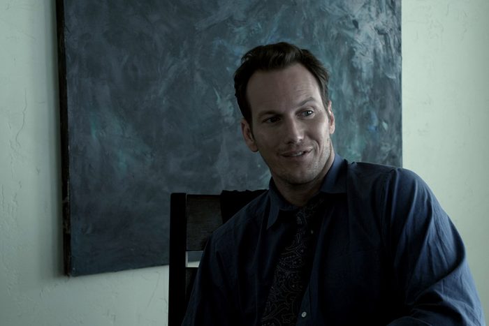 Man smiling, sitting in a chair, against a dark abstract painting on a light-colored wall.