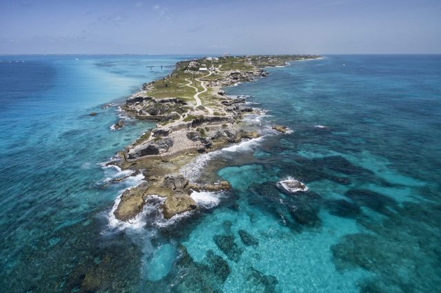 Rocky coast stretches into turquoise ocean; clear sky above; winding path follows the shoreline, surrounded by sparse vegetation and distant structures.