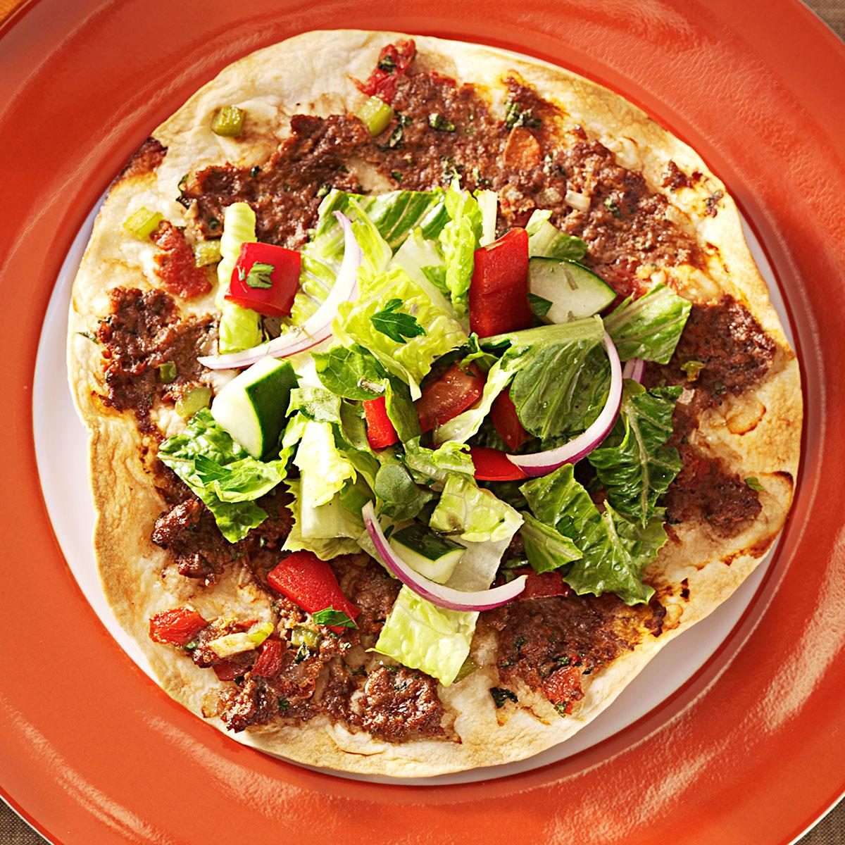 Flatbread holds seasoned meat and fresh salad, served on an orange plate in a dining setting.