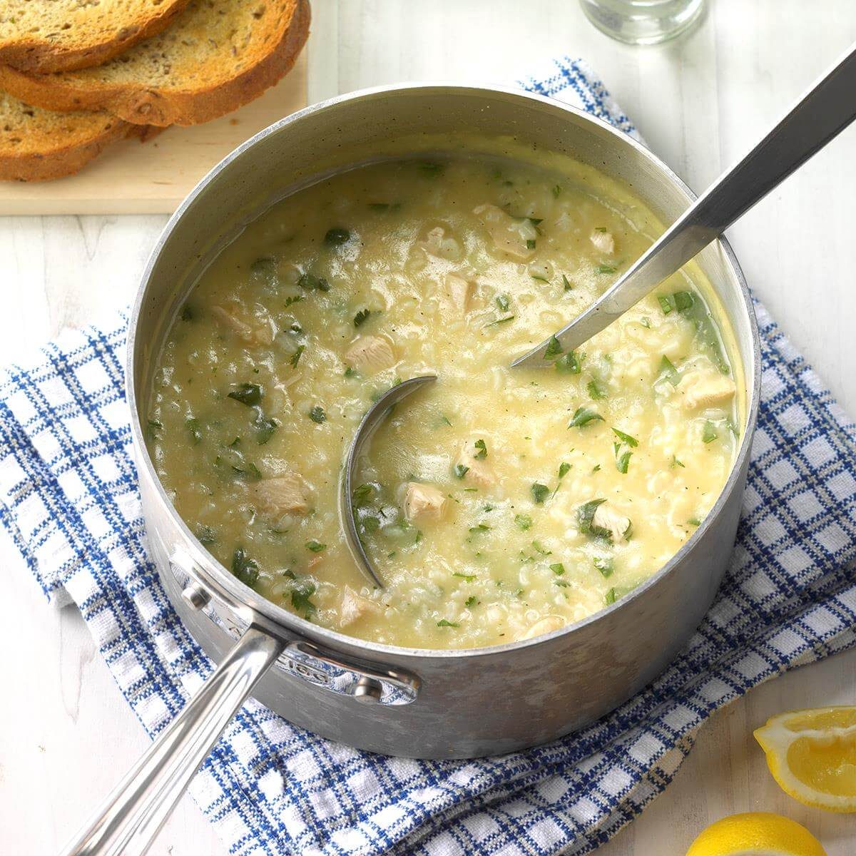 Soup simmers in a pot with a ladle, resting on a blue checkered cloth, surrounded by bread slices and lemon wedges.