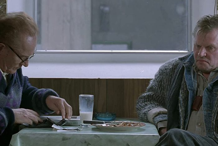 Two men sit at a table; one reads a newspaper, the other looks forward. A window and empty plates are in the background.