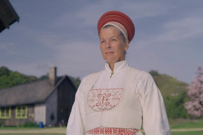 A woman stands wearing traditional attire with red embroidery, in a rural setting with a wooden building and flowering tree in the background.
