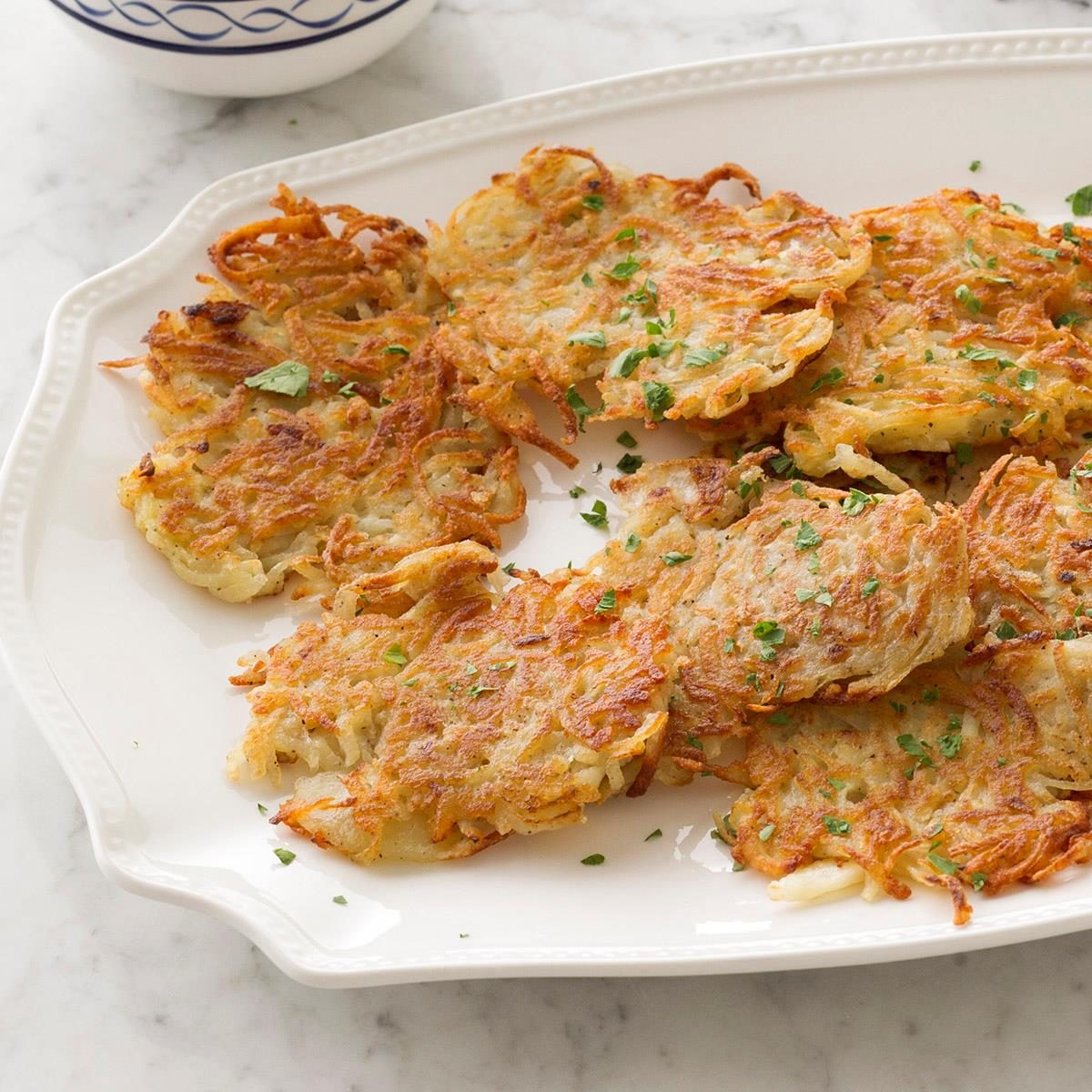 Latkes are arranged on a decorative white platter, garnished with chopped herbs, resting on a marble surface.