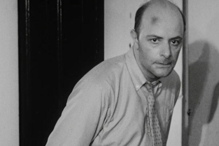 A man with a serious expression leans forward in a dimly lit room, wearing a shirt and tie.