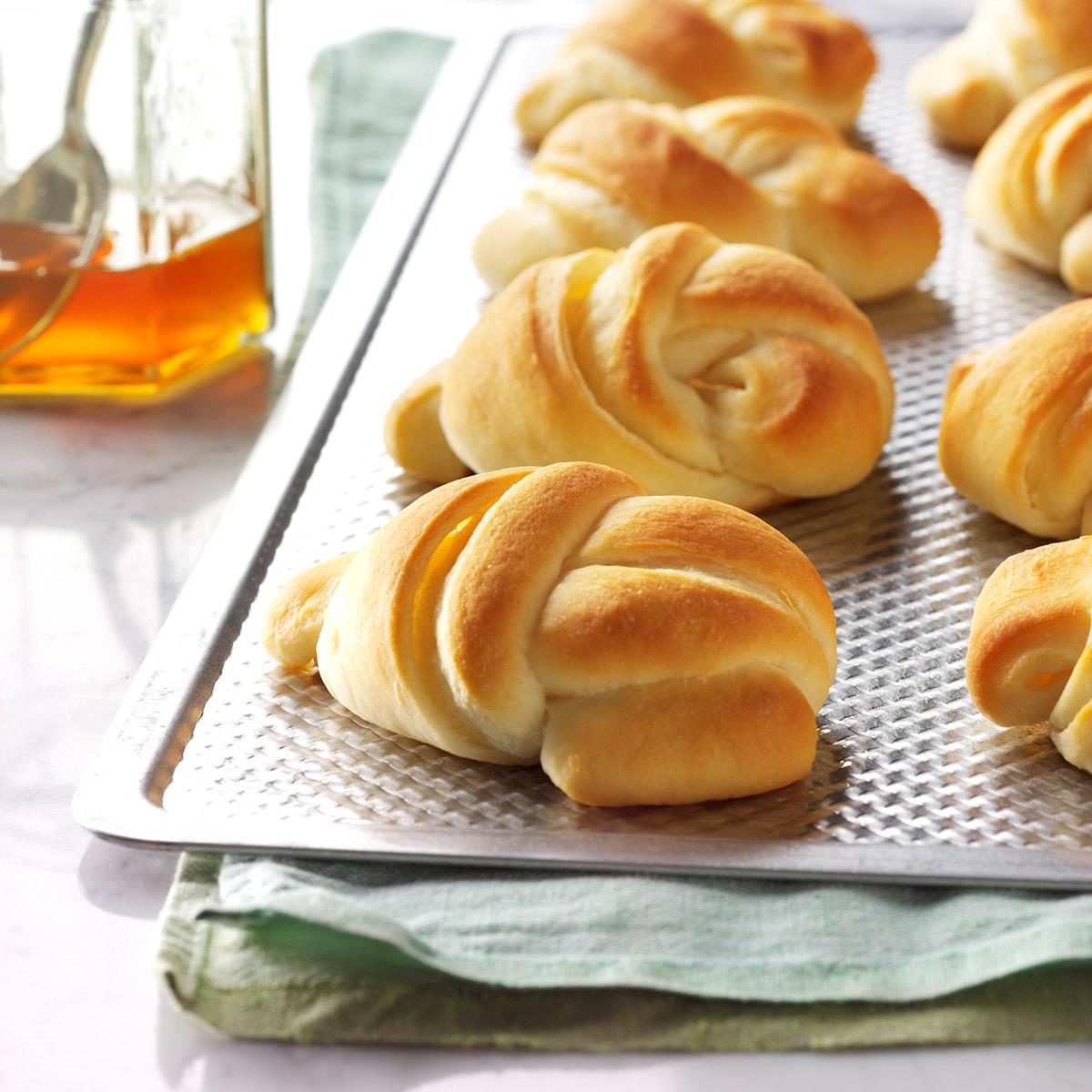Golden brown dinner rolls sit on a textured baking sheet, with a jar of honey and spoon nearby on a white table.