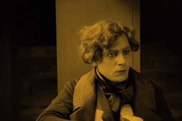 Person sits, looking pensive, wearing a scarf and coat; dimly lit, sepia-toned setting with a plain background, creating a vintage atmosphere.