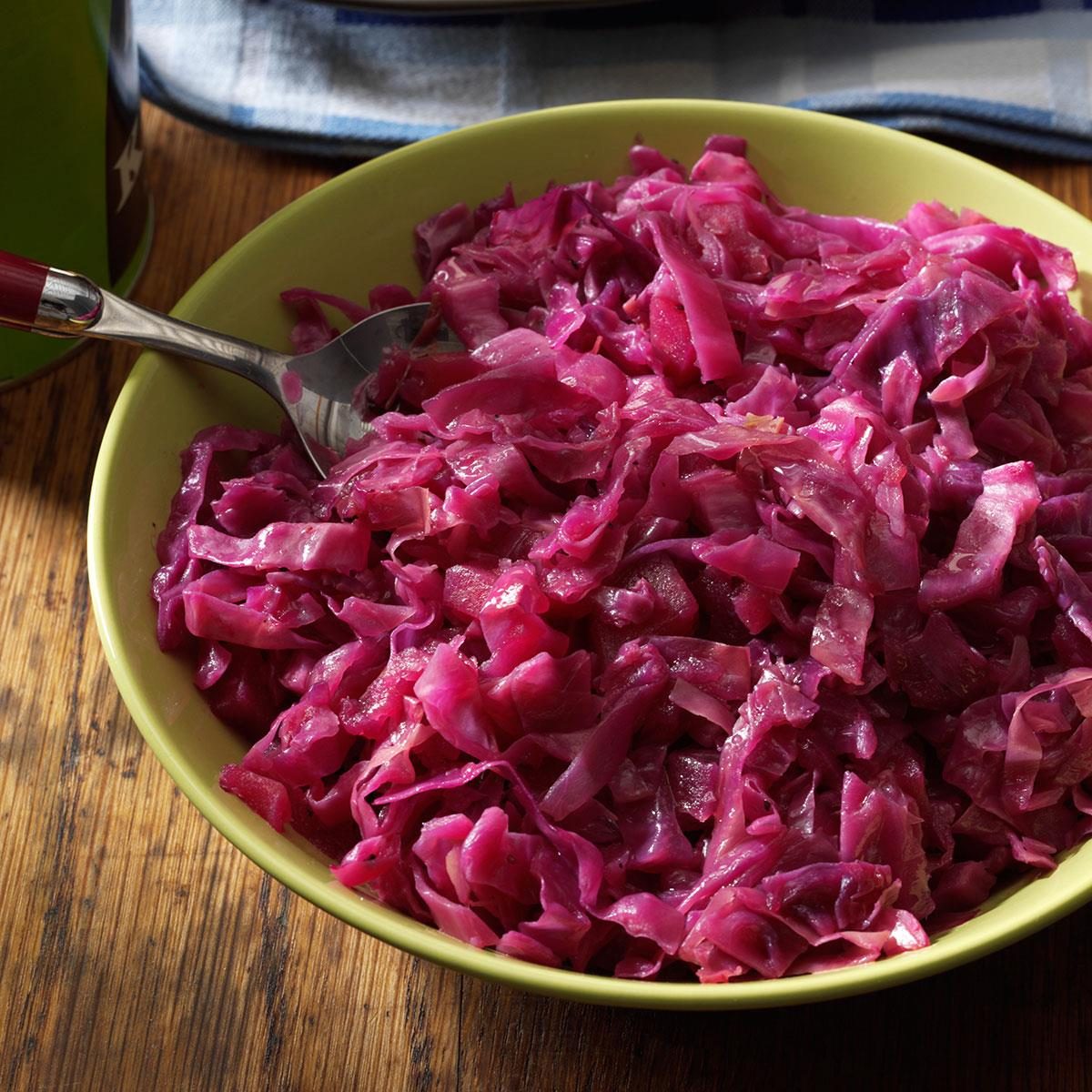 Purple cabbage fills a green bowl, accompanied by a serving fork, on a wooden table with a cloth nearby.