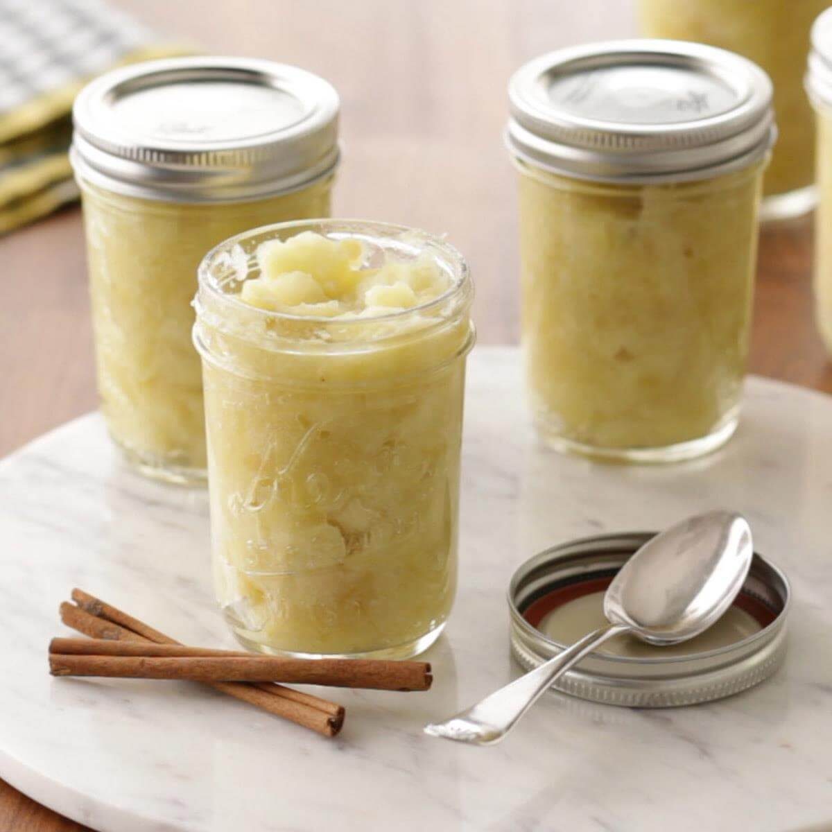 Jars filled with applesauce sit on a marble surface. A spoon rests on a lid nearby, and cinnamon sticks lie beside it.