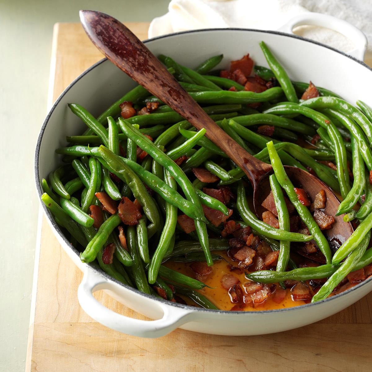 Green beans mixed with bacon are being stirred with a wooden spoon in a white pot on a wooden surface.