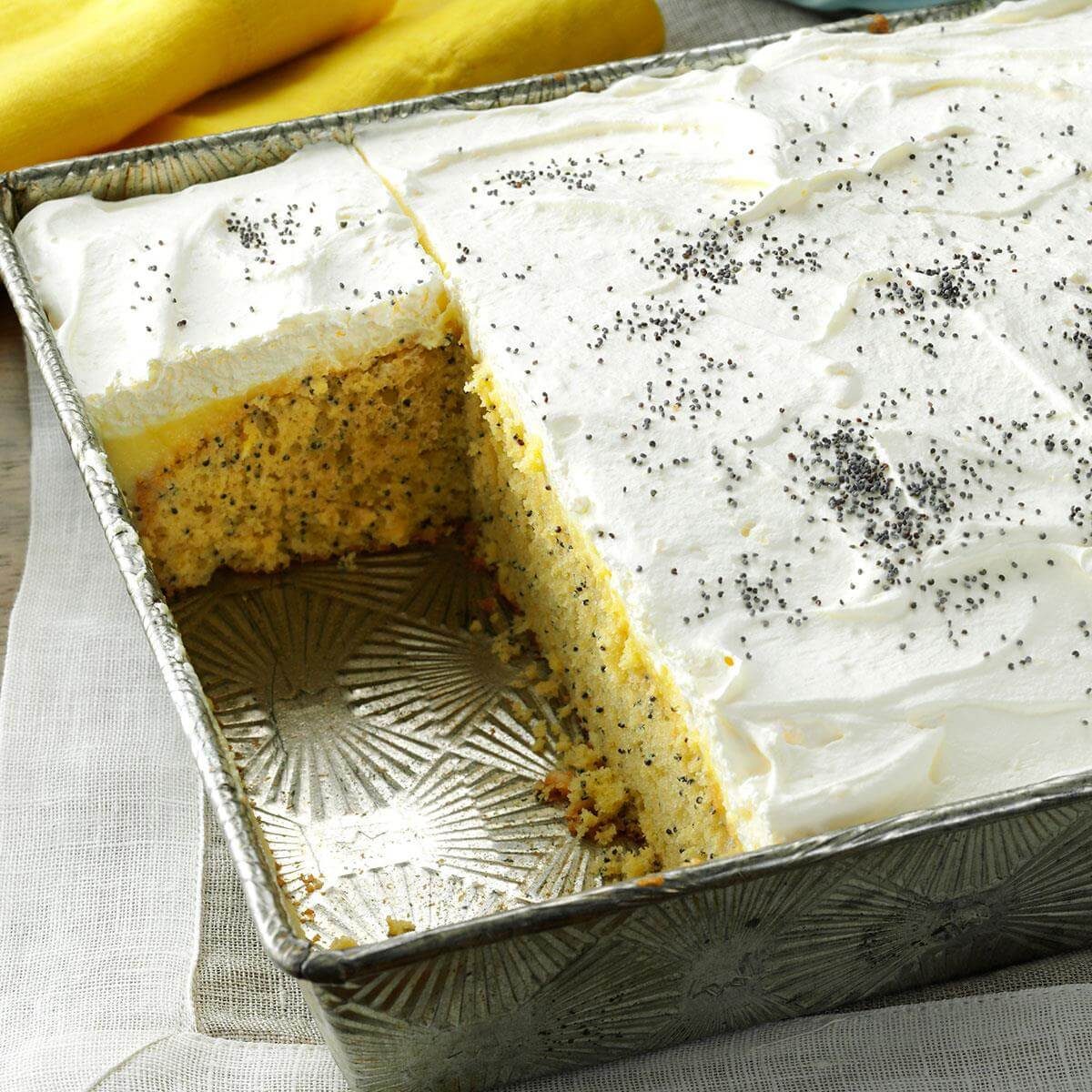 Cake topped with frosting and poppy seeds, partially sliced in a metal baking pan on a fabric napkin, with yellow cloth background.