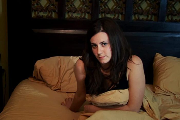 A woman sits on a bed, partially covered by a beige blanket, against a dark wooden headboard with ornamental design.