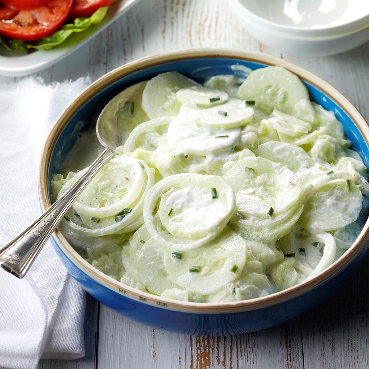 Cucumber slices sit in creamy dressing in a blue bowl, with a spoon, on a wooden table, next to sliced tomatoes and lettuce.