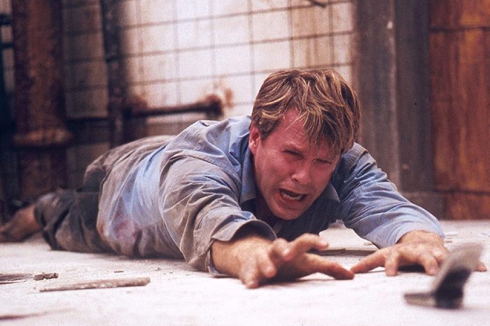 A desperate man crawls on the ground, reaching toward an object, in a grimy, industrial-tiled room.