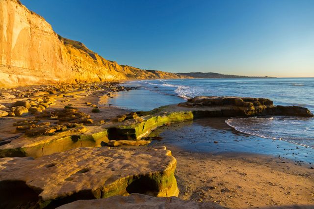 Rocks bathed in golden sunlight sit along a beach with cliffs and waves under a clear blue sky.