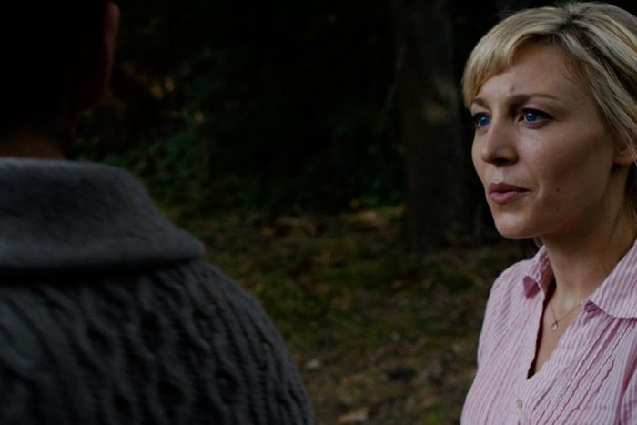 A person converses outdoors, wearing a pink striped shirt, with trees and greenery in the background. Another individual is partially visible.