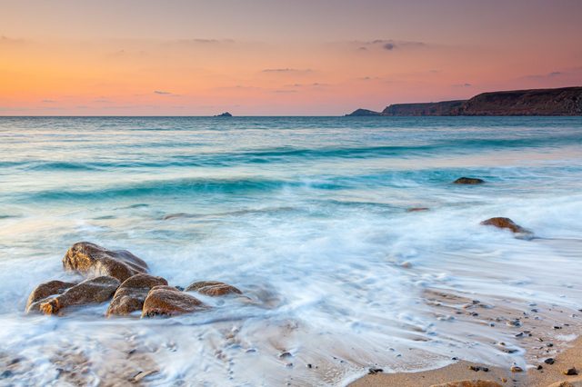 Waves gently wash over rocks on a sandy beach at sunset, with distant cliffs under a colorful sky.